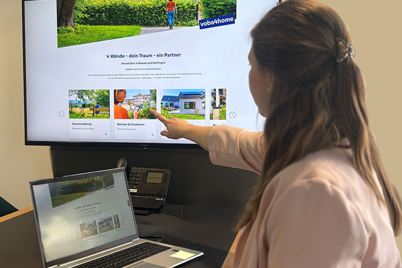 Frau sitzt an einem Schreibtisch und zeigt mit der Hand auf einen Fernseher, auf dem das Thema „Rechner und Checklisten“ ange