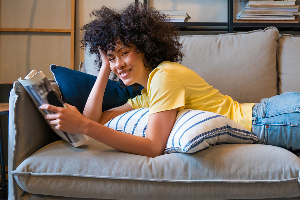 Mädchen mit dunklen Locken liegt auf Sofa mit Zeitschrift in der Hand