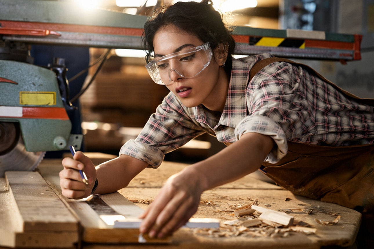 Eine junge Frau trägt eine Schutzbrille und arbeitet in einer Werkstatt mit Holz.