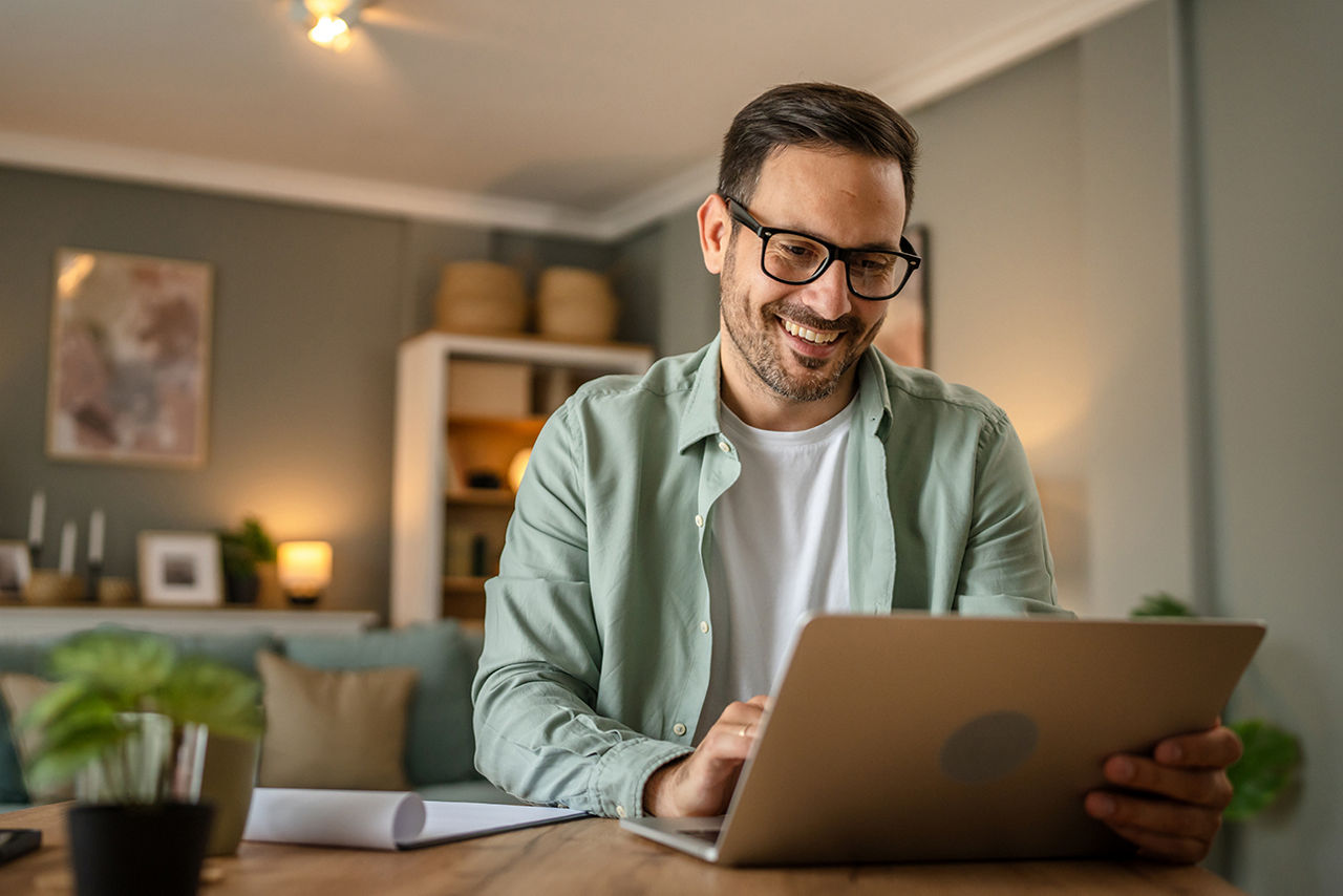 Mann mit Brille sitzt lächelnd am Schreibtisch und schaut auf seinen Laptop