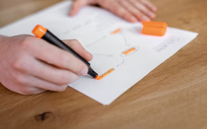 Eine Person nutzt einen orangefarbenen Marker, um auf einem weißen Blatt Papier ein Flussdiagramm zu zeichnen.