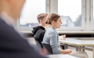 Das Bild zeigt ein Klassenzimmer, in dem eine junge Frau aufmerksam am Schreibtisch sitzt und nach vorne blickt.