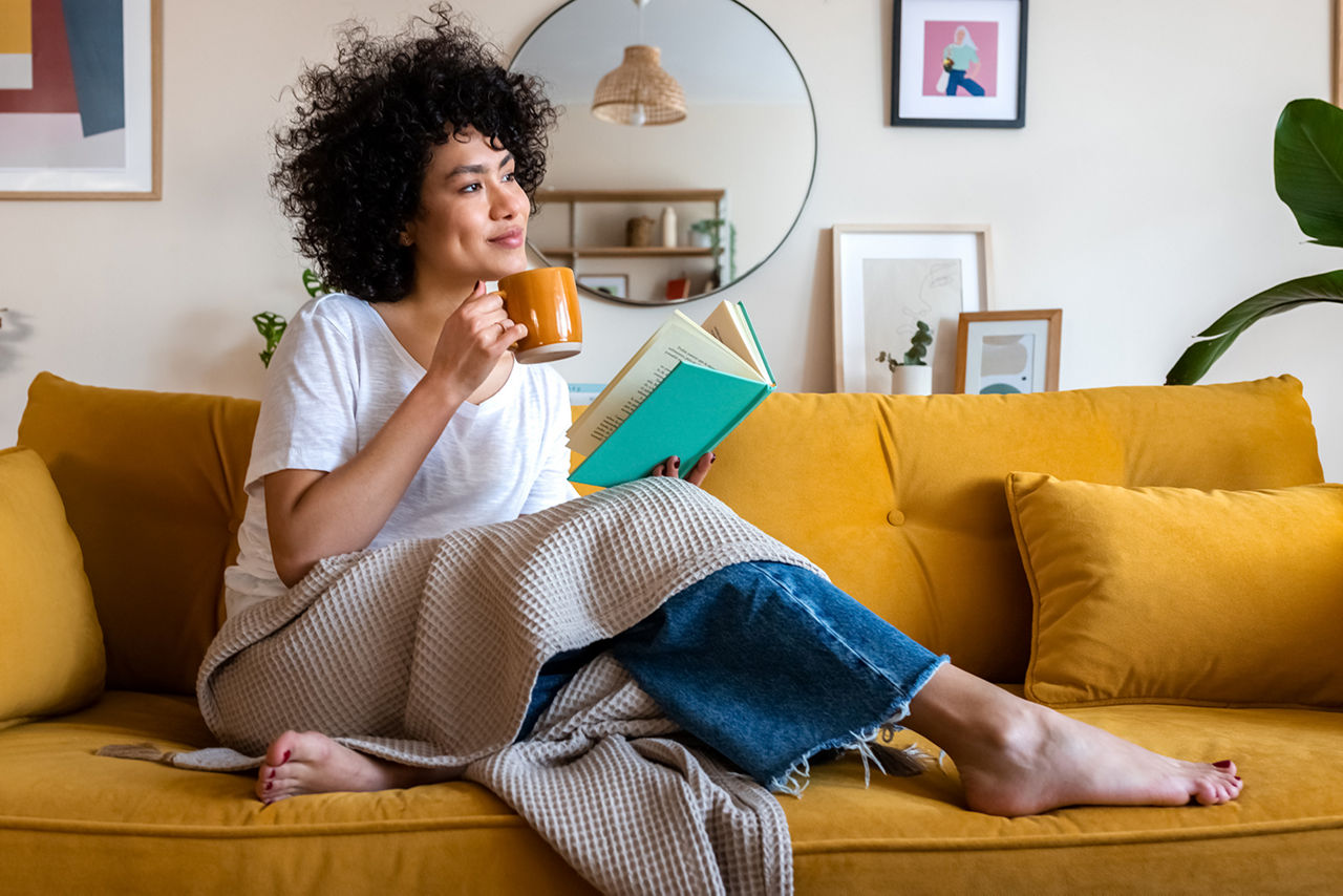 Nachdenkliche, entspannte Afroamerikanerin liest zu Hause ein Buch und trinkt Kaffee auf der Couch