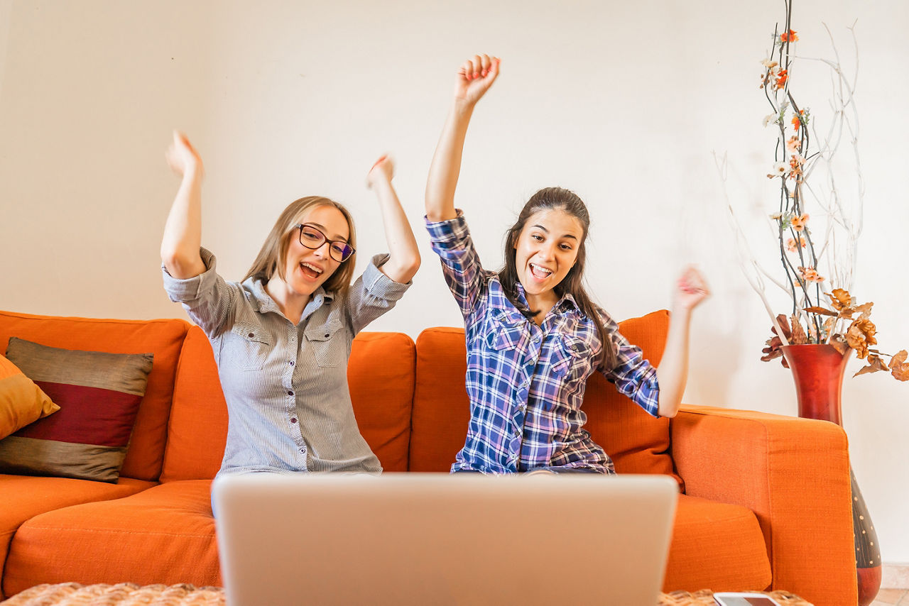Zwei Frauen sitzen nebeneinander auf einem Sofa mit Blick auf einen Laptop und freuen sich