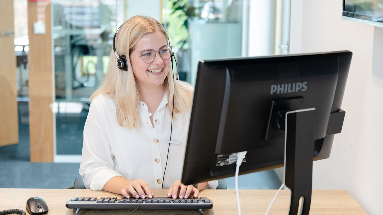 Eine junge Frau mit blondem Haar und Brille sitzt lächelnd vor einem Computer und trägt ein Headset