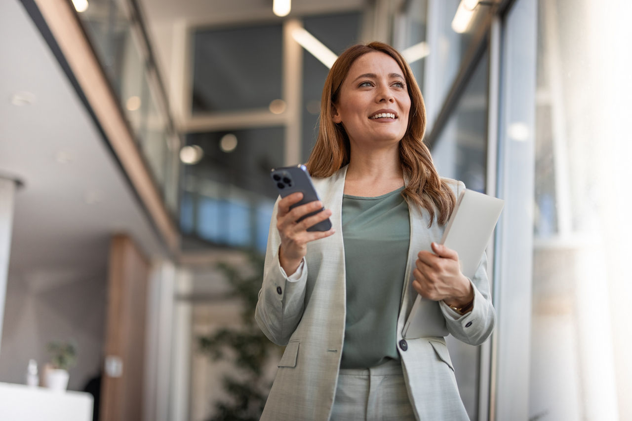 Eine Frau steht im Office und hält ihr Handy und Unterlagen in den Händen