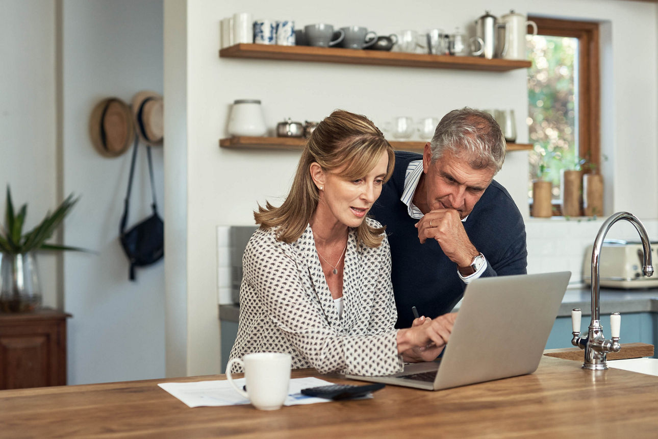 Ein Mann und eine Frau sitzen am Küchentisch und schauen in einen Laptop.