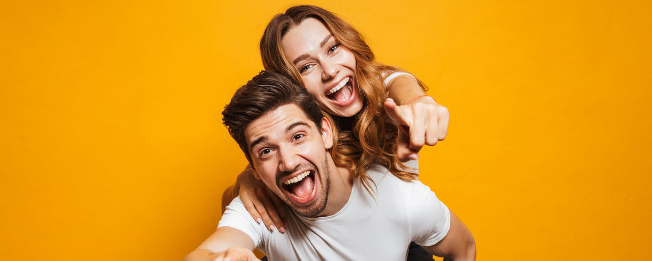 Image of caucasian man giving piggyback ride to joyful woman and pointing fingers at you isolated over yellow background
