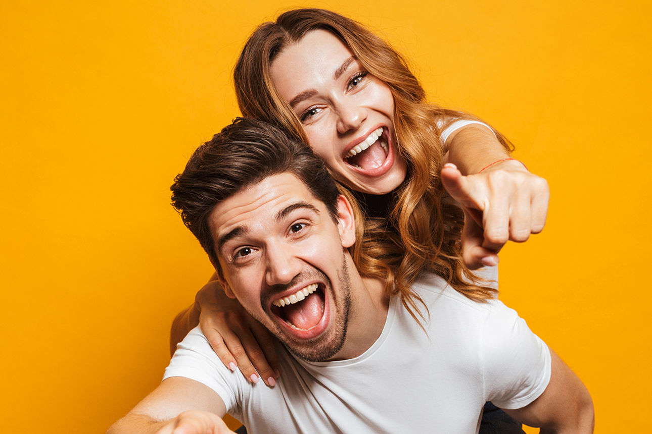 Image of caucasian man giving piggyback ride to joyful woman and pointing fingers at you isolated over yellow background