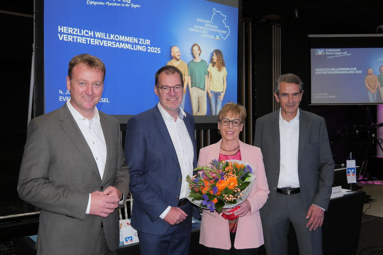 Drei Herren und eine Dame mit einem Blumenstrauß in der Veranstaltungshalle und hinter Ihnen zwei Monitore. 