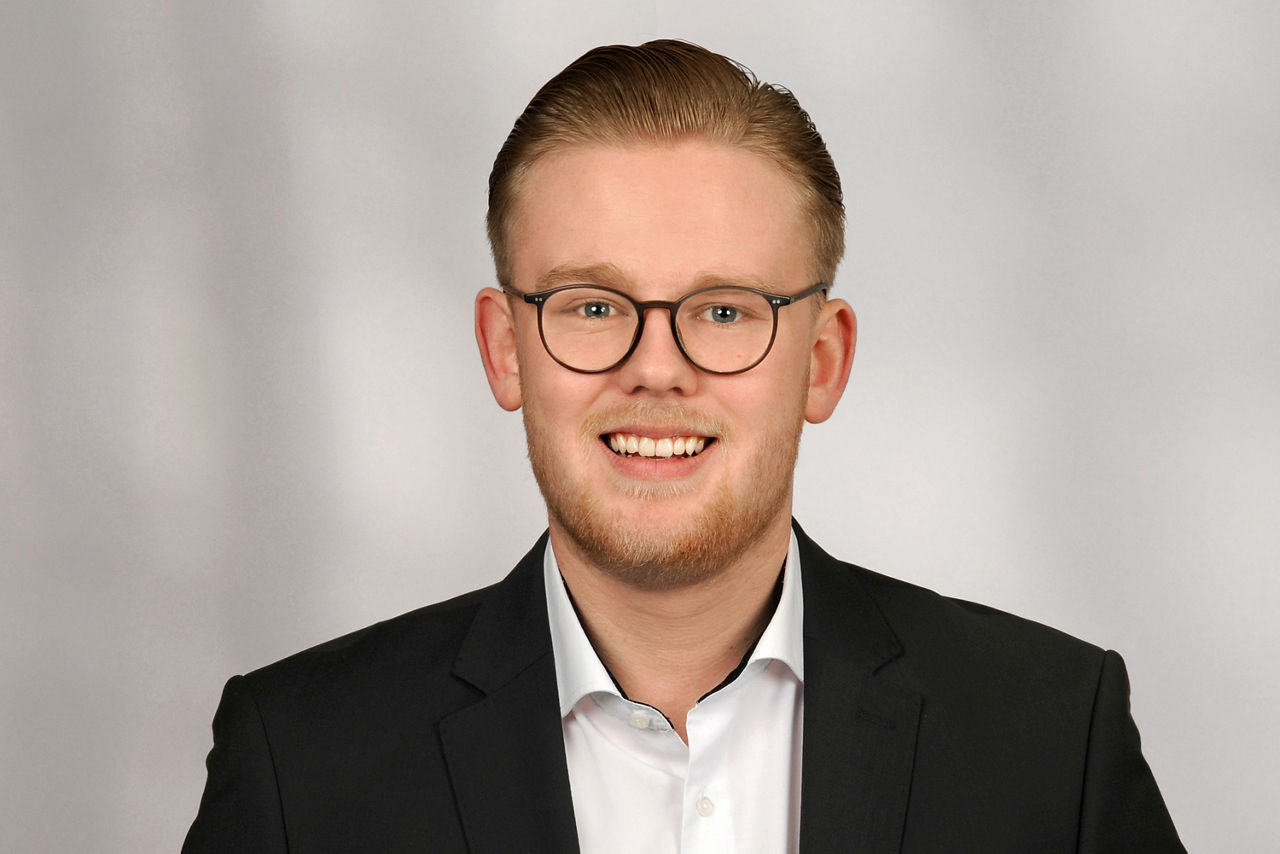 Mann mit kurzen, zurückgekämmten blonden Haaren trägt schwarzen Anzug und weißes Hemd vor neutralem Hintergrund