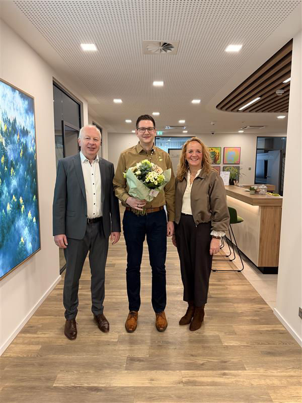 Zwei Männer und eine Frau haben sich zum Foto aufgestellt. Der Mann in der Mitte hat einen Blumenstrauß in der Hand.