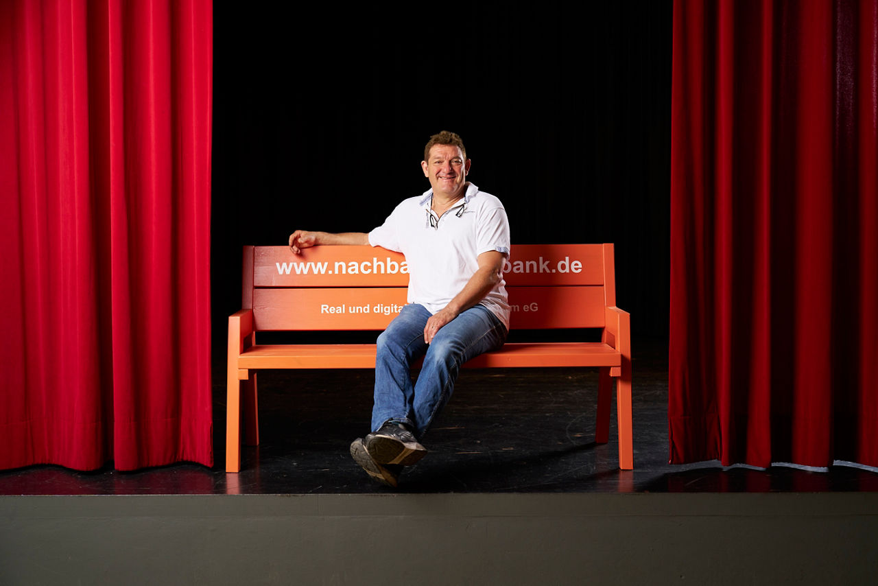 Thomas Boxhammer sitzt auf einer orangen Bank auf einer Bühne mit roten Vorhängen