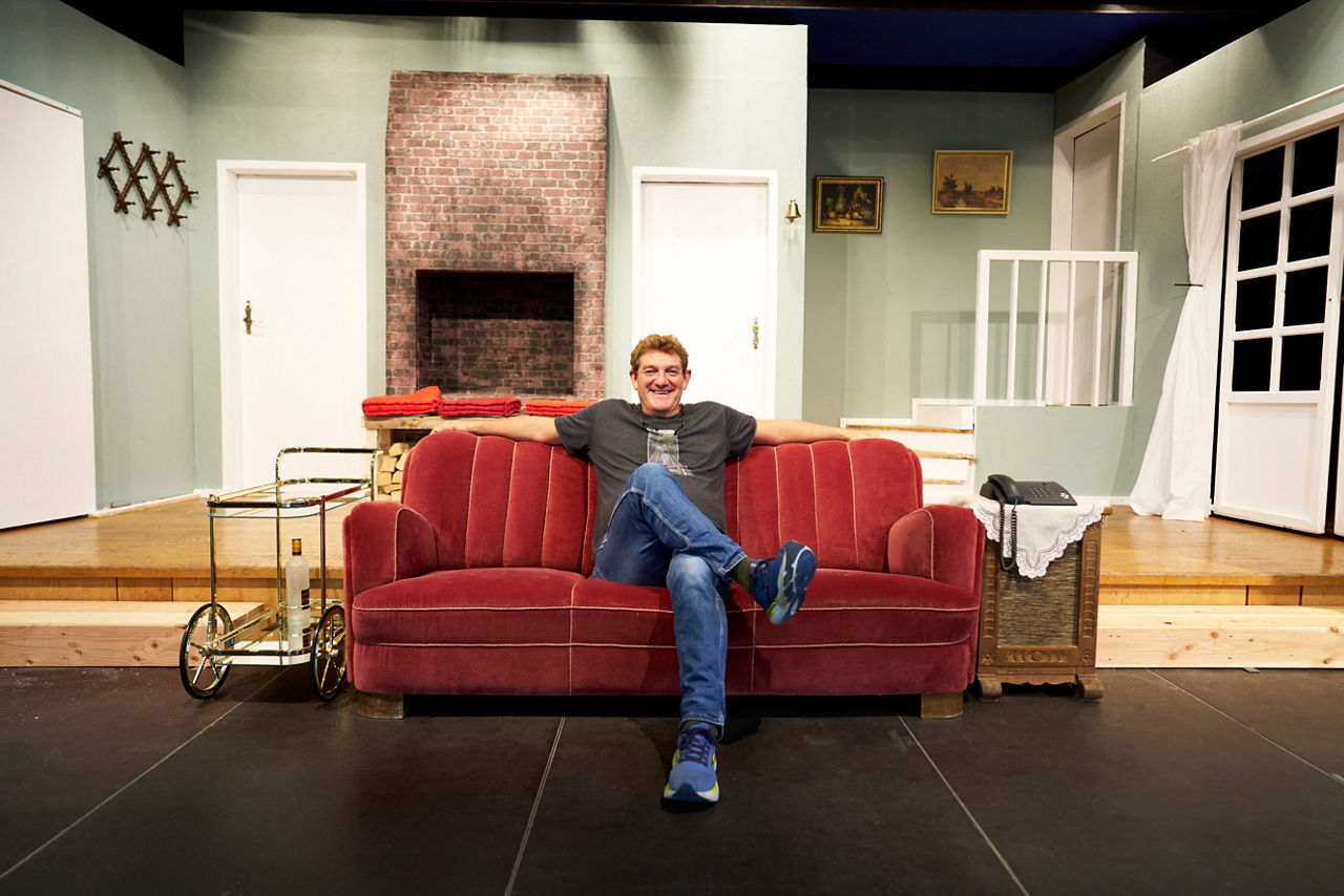 Thomas Boxhammer sitzt auf einem roten Sofa vor einer Kulisse