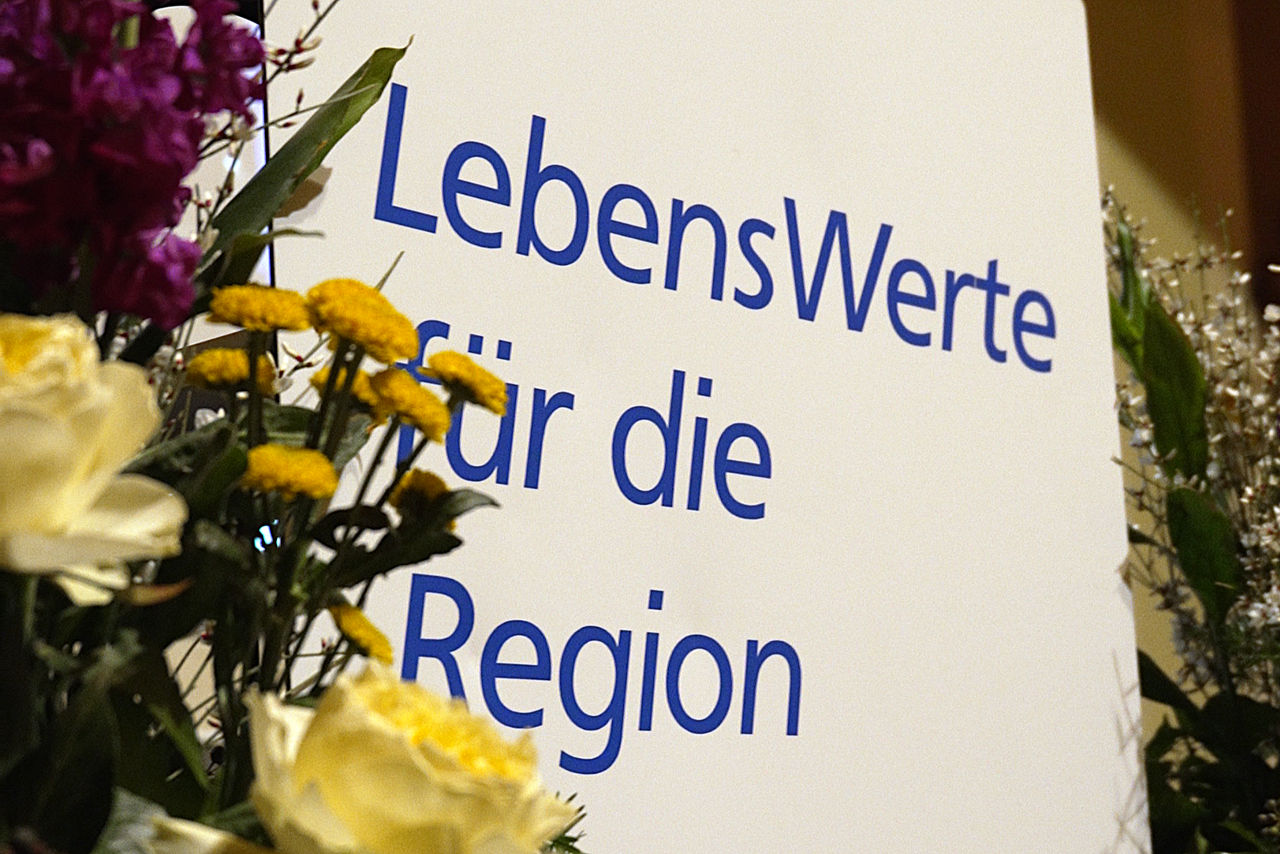 Foto einer Leinwand-Projektion mit dem Schriftzug "LebensWerte für die Region", im Vordergrund Blumen-Dekoration