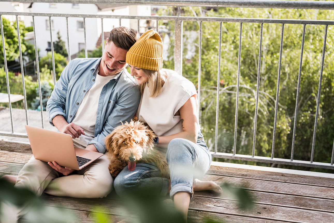 EIn junges Paar dass sich am Balkongeländer anlehnt und in einen Laptop schaut