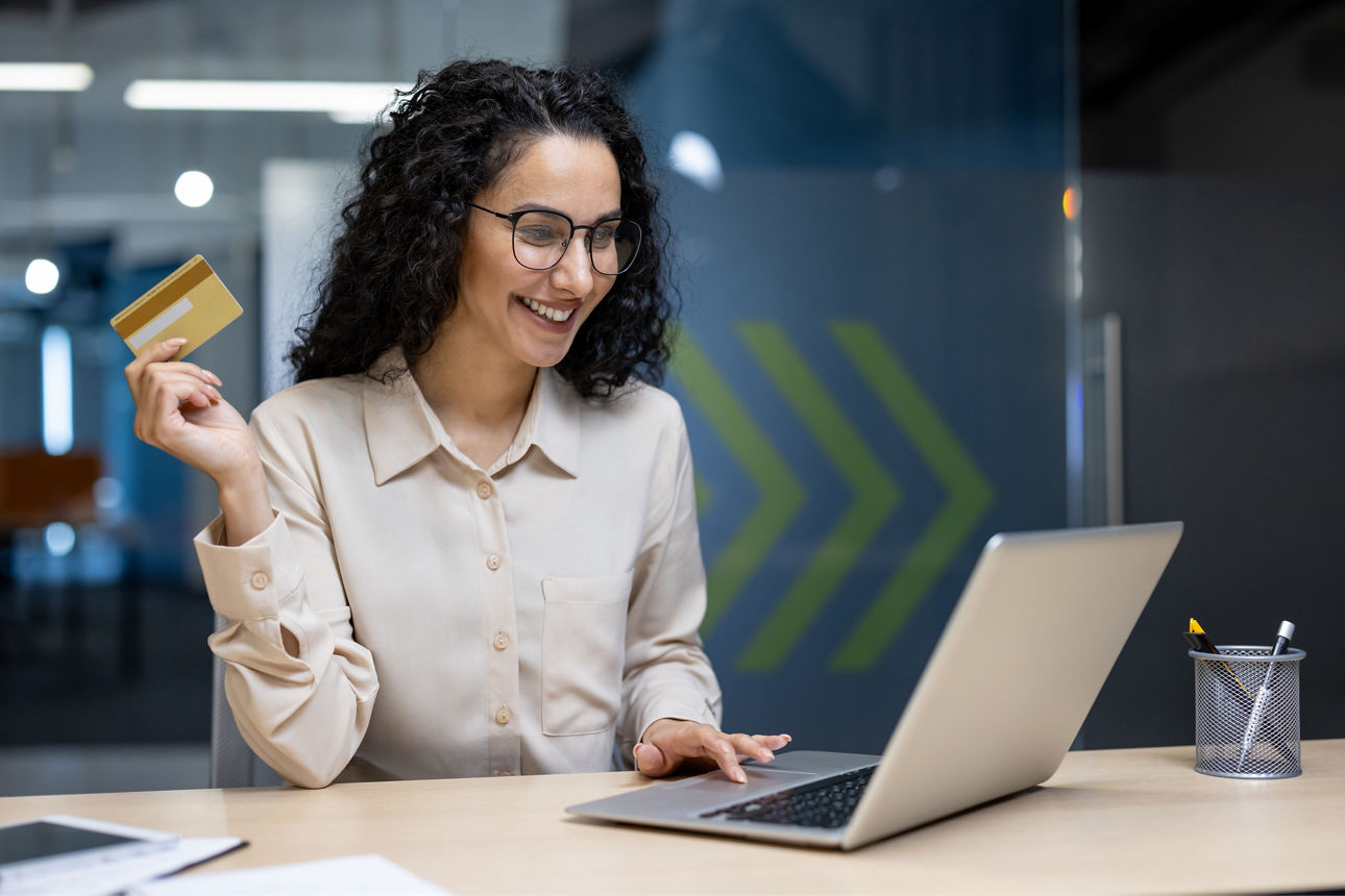 Eine junge Frau mit guter Laune sitzt mit ihrer girocard am Schreibtisch und schaut auf einen Laptop