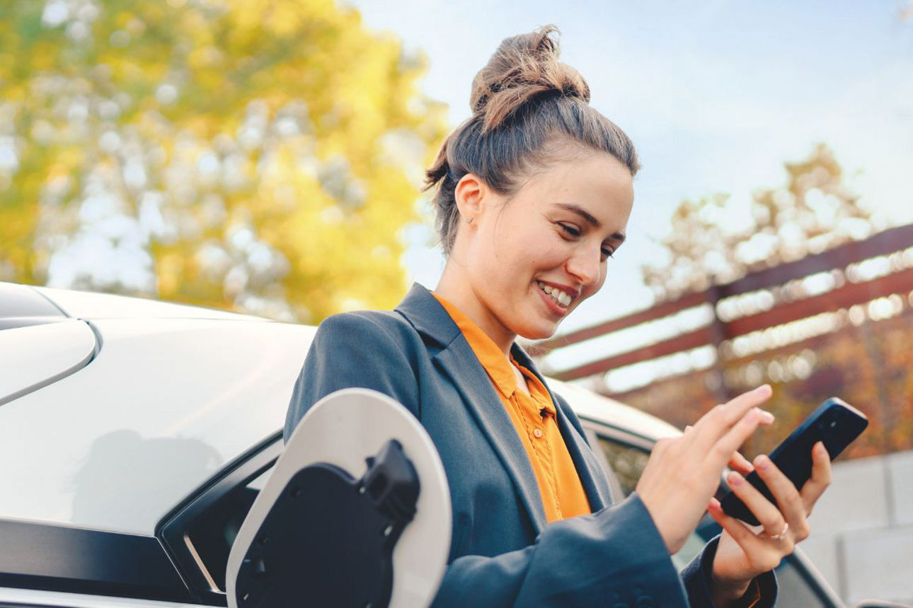 Frau lehnt an einem Elektroautu und schaut in ihr Smartphone
