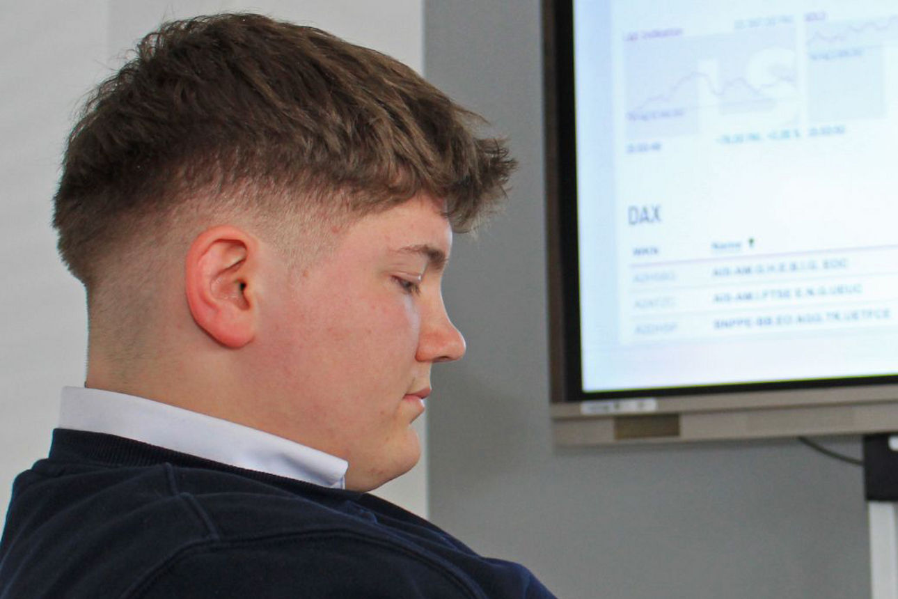 Junger Mann sitzt vor Börsendaten auf einem großen Bildschirm und schaut auf sein Tablet