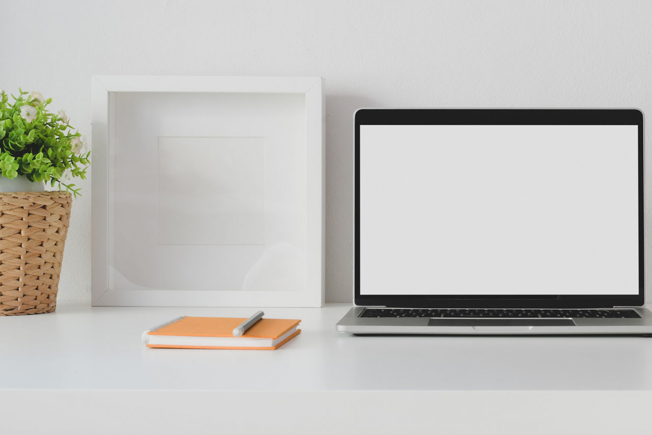 Schreibtisch auf dem eine Pflanze, ein Bilderrahmen, ein Block und Stift und ein Laptop mit WhiteScreen stehen 