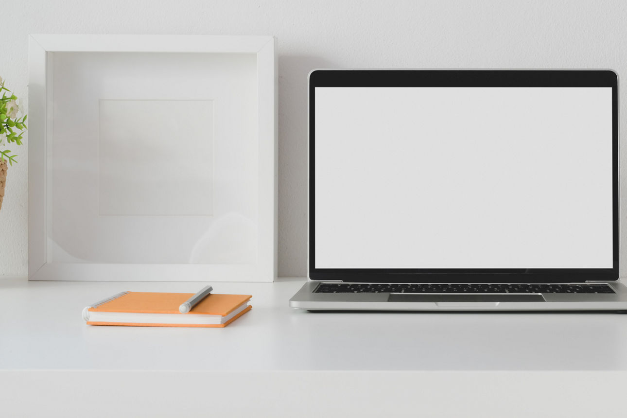 Schreibtisch auf dem eine Pflanze, ein Bilderrahmen, ein Block und Stift und ein Laptop mit WhiteScreen stehen 