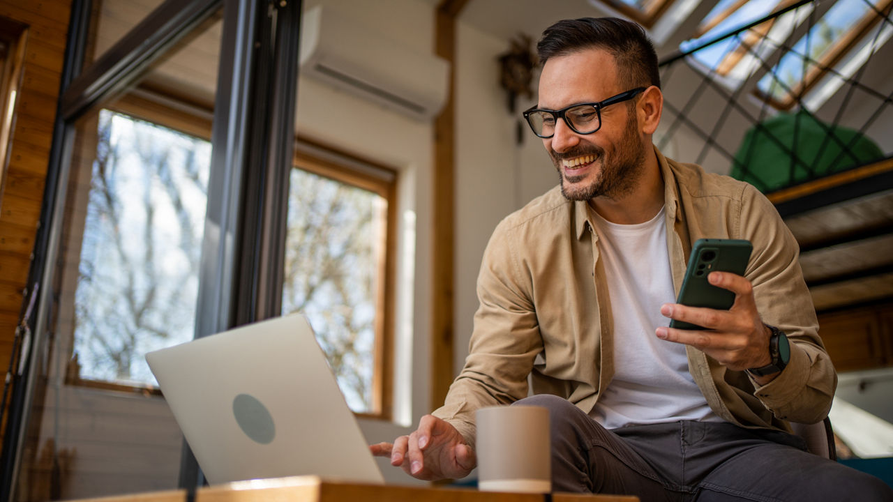 TraderDepot - Ein junger Mann nutzt lächelnd seinen Computer und sein Handy