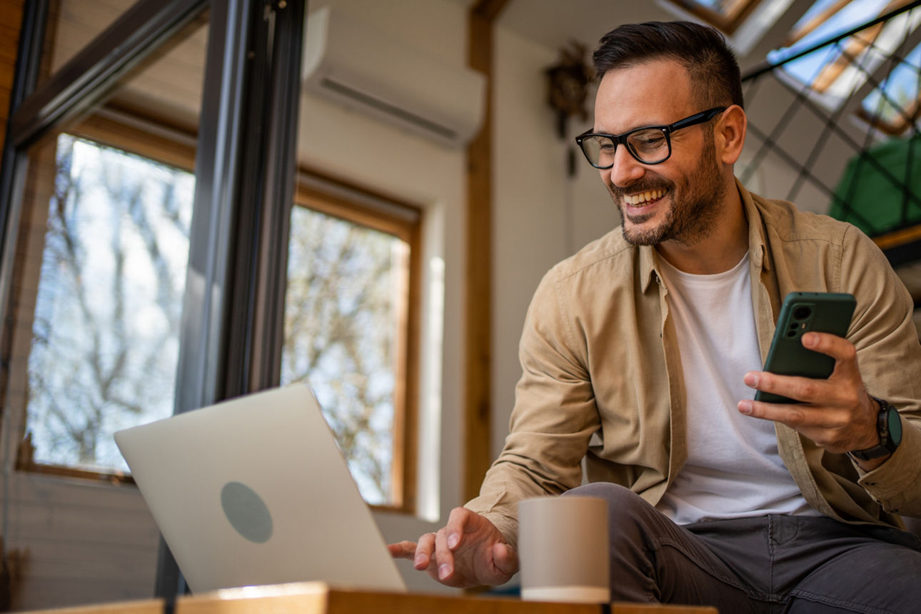 TraderDepot - Ein junger Mann nutzt lächelnd seinen Computer und sein Handy