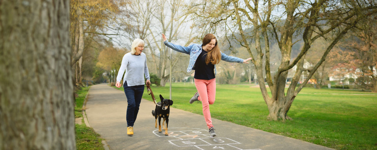 Eine Frau hüpft im Park auf einen Himmel & Hölle Spielfeld. Daneben läuft eine älter Frau mit ihrem Hund.