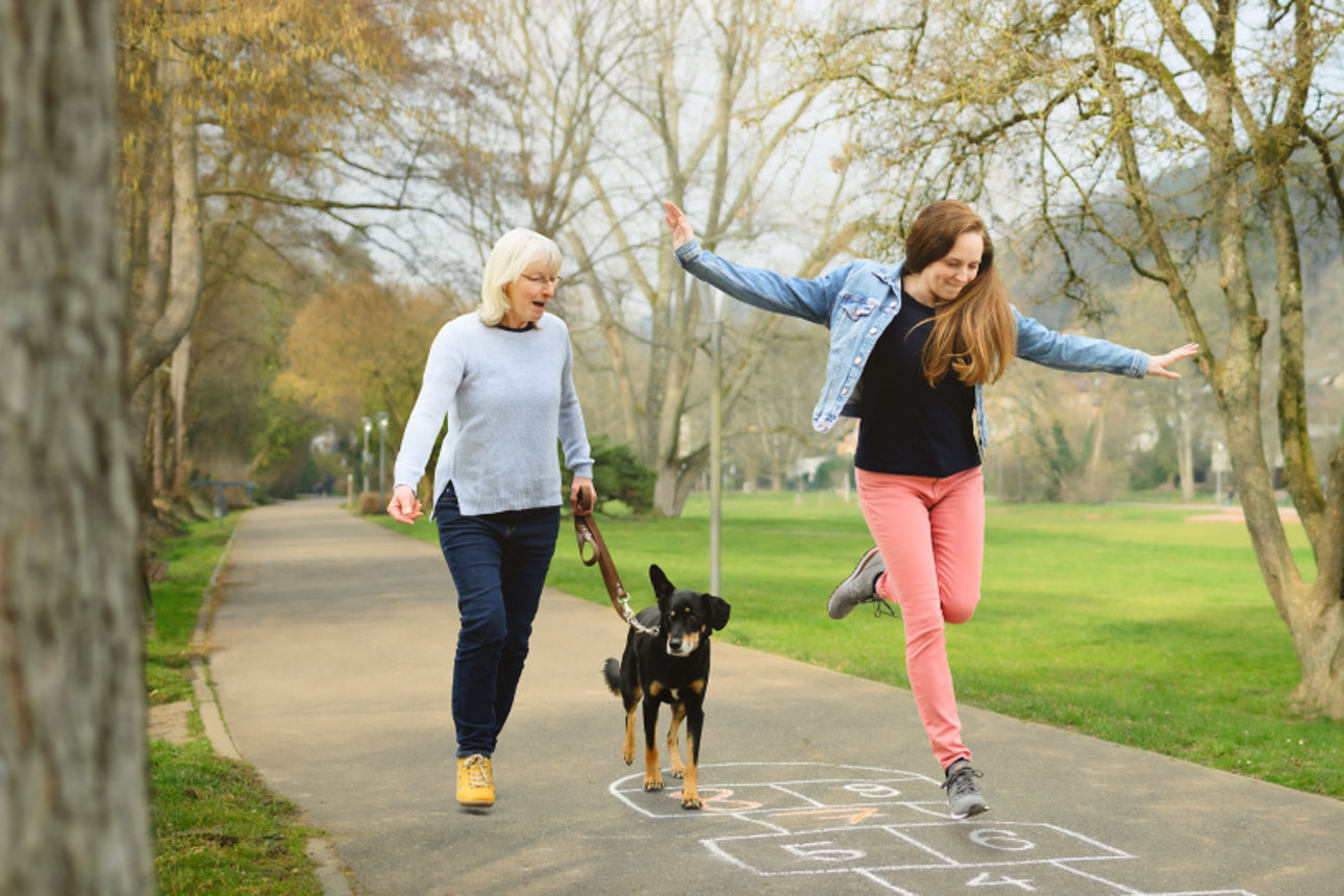 Eine Frau spaziert mit einem Hund, während ein Mädchen auf einem Gehweg hüpft und auf ein Hüpfspiel aus Kreide springt.