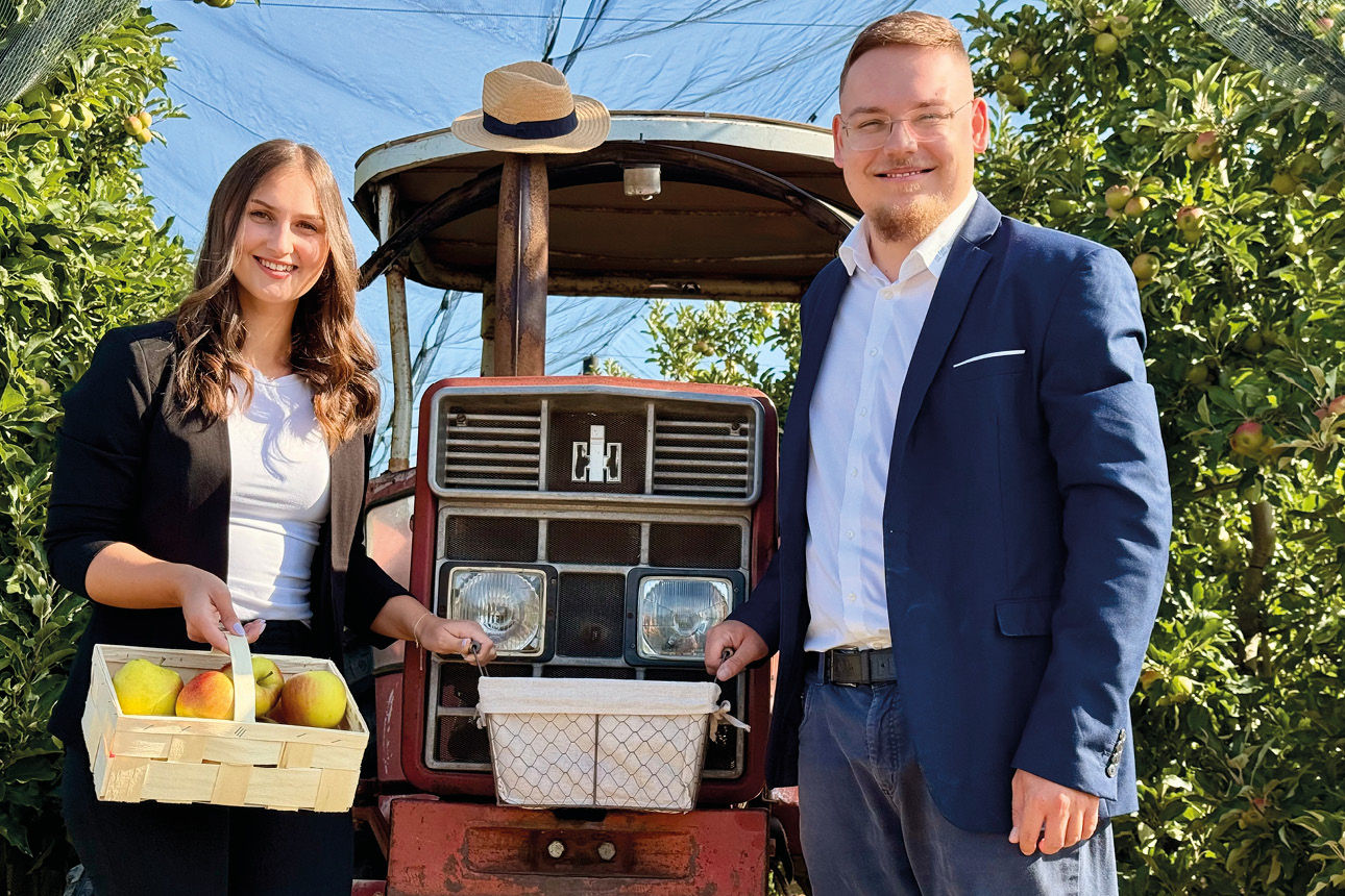 Auf dem Bild sehen Sie zwei Personen, die auf einer Apfelplantage stehen