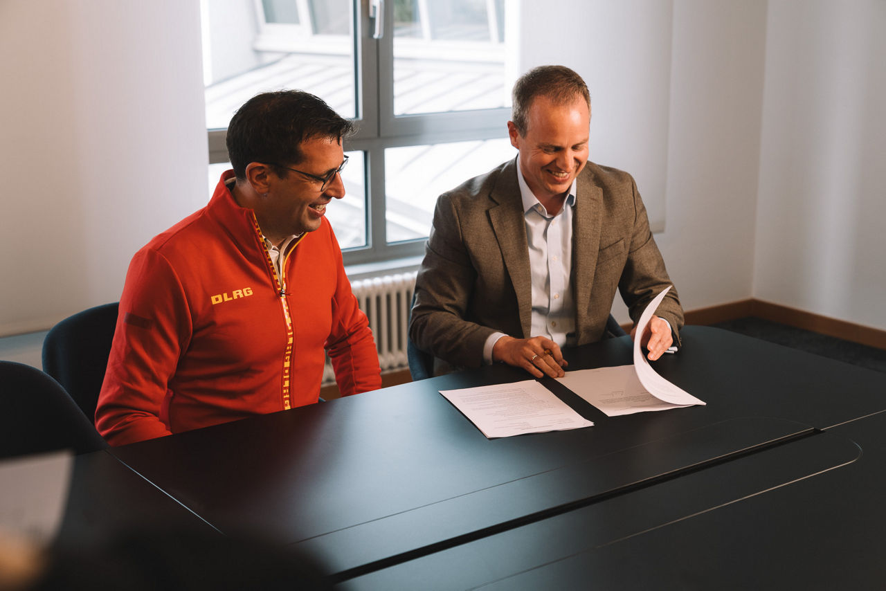 Zwei Personen an einem Konferenztisch beim Unterschreiben von Dokumenten, Fenster im Hintergrund.
