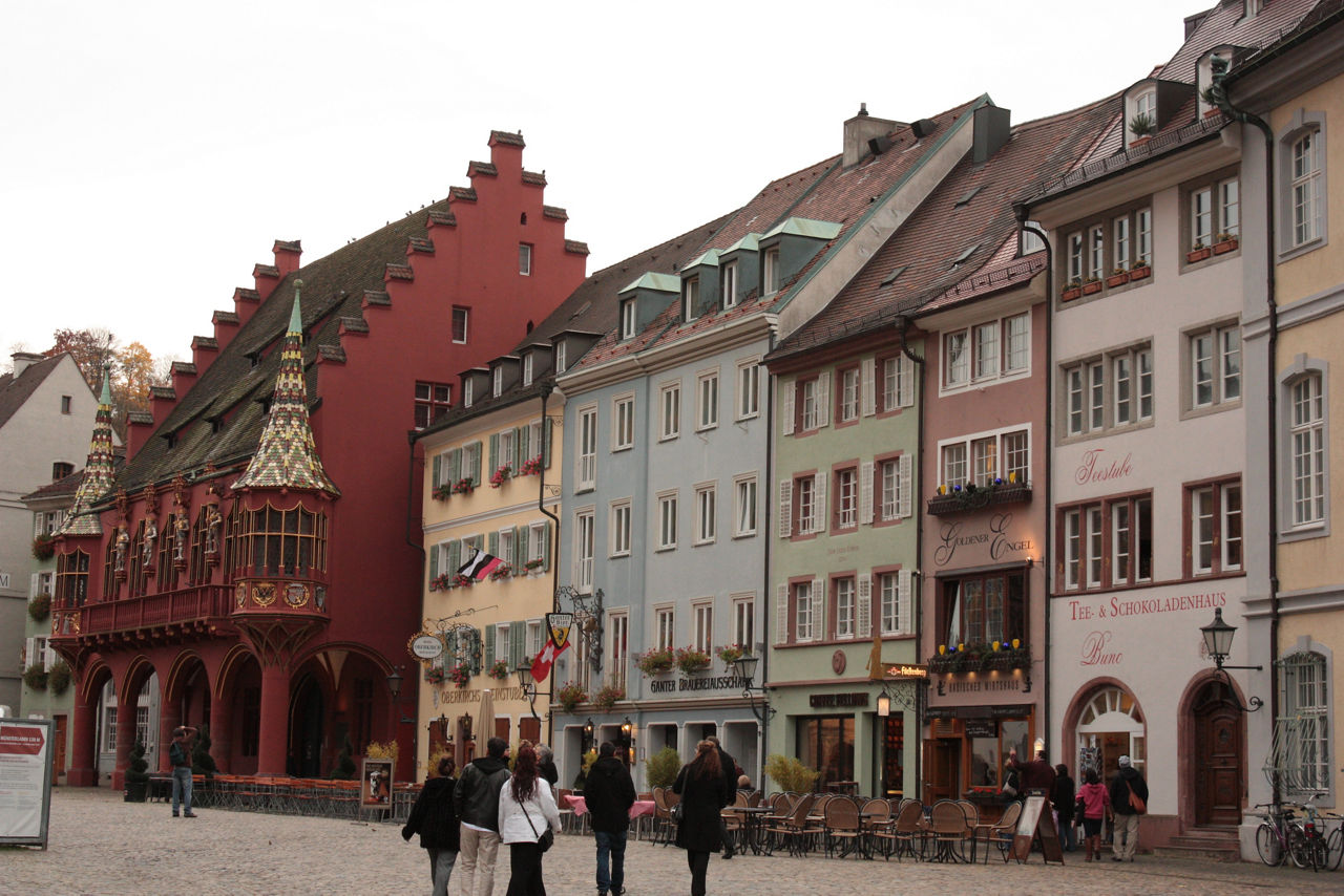 Das Bild zeigt eine Reihe farbenfroher, mehrstöckiger Altstadthäuser mit traditionellen Fassaden, Erkern und einem auffällig roten Gebäude mit Ziergiebeln und Türmchen. Vor den Häusern befinden sich Straßencafés, Geschäfte und mehrere Passanten auf dem Kopfsteinpflaster.