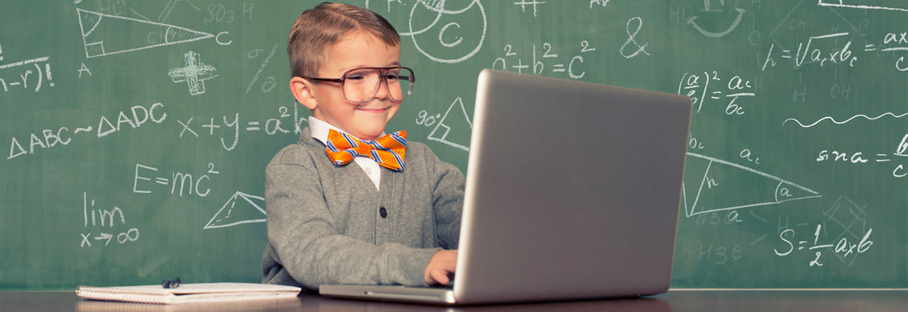 Bild eines kleinen Jungen mit einer großen Brille, orangenen Fliege und Laptop vor einer beschrifteten Schultafel.