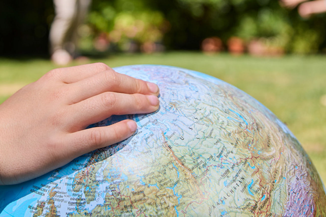 Eine Kinderhand liegt auf einem Globus im Garten. Ein grüner Punkt mit Text: Ein Fonds exklusiv für unsere Kunden!