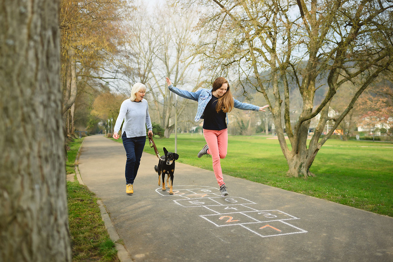 Eine Frau spaziert mit einem Hund, während ein Mädchen auf einem Gehweg hüpft und auf ein Hüpfspiel aus Kreide springt