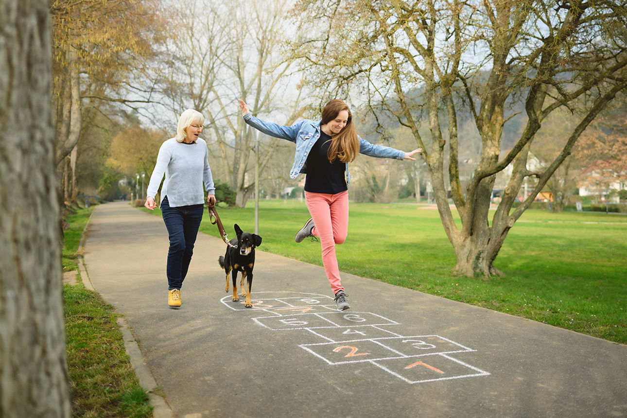 Eine Frau spaziert mit einem Hund, während ein Mädchen auf einem Gehweg hüpft und auf ein Hüpfspiel aus Kreide springt