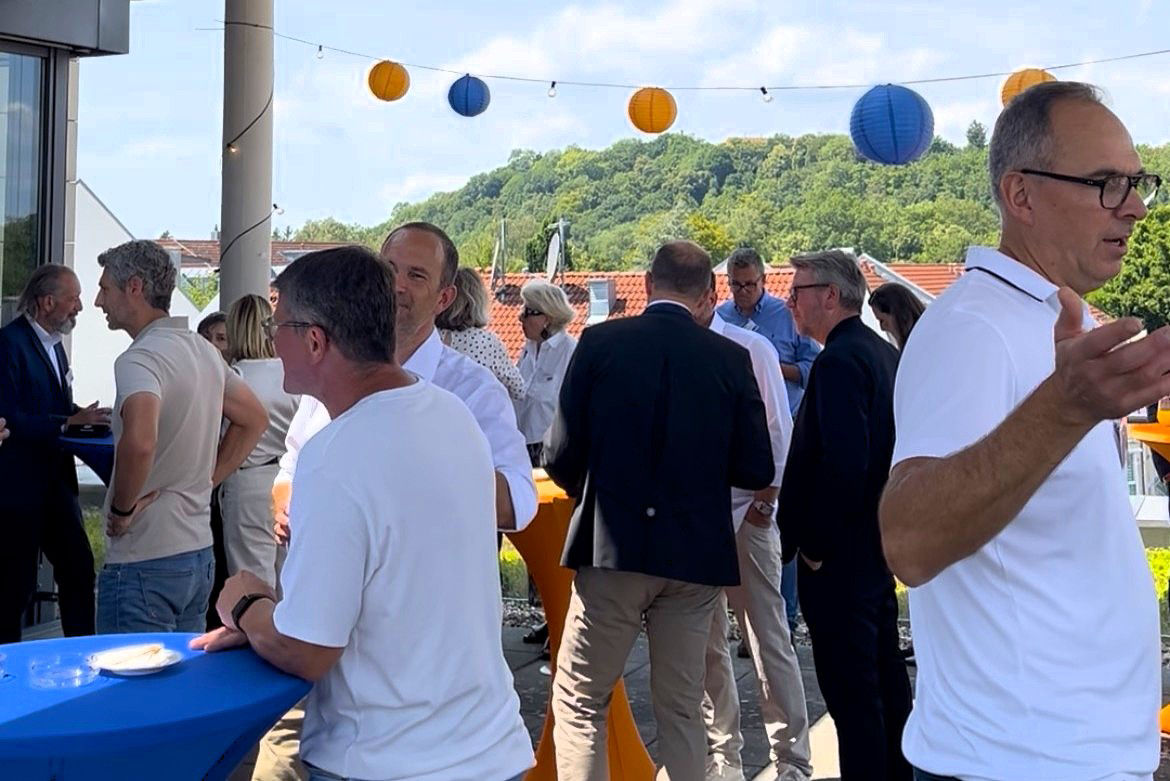 Eine gemischte Gruppe von Personen stehen sich unterhaltend auf einer Dachterrasse. Im Hintergrund grüne Hügel