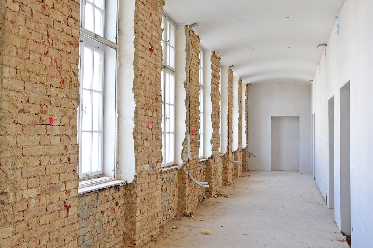 Langer, schmaler Flur in einem Gebäude während der Renovierung. Links freigelegtes Ziegelmauerwerk mit hohen Fenstern, rechts weiß verputzte Wand mit Türen. Der Boden ist uneben, Kabel hängen lose von der Wand.