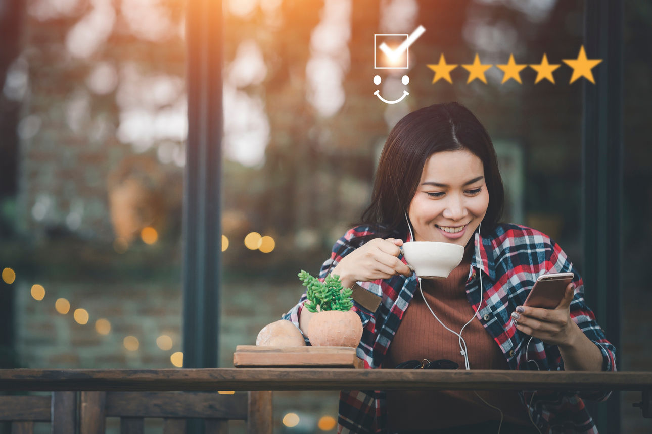 Frau sitzt draußen an einem Tisch mit einem Getränk und  macht eine online Umfrage am Handy, darüber sind 5 Sterne als Bewertung über ihren Kopf zu sehen.