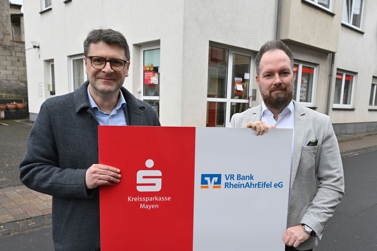 Vertreter der Kreissparkasse Mayen und VR Bank RheinAhrEifel eG präsentieren gemeinsames Schild vor Sparkassen-Filiale