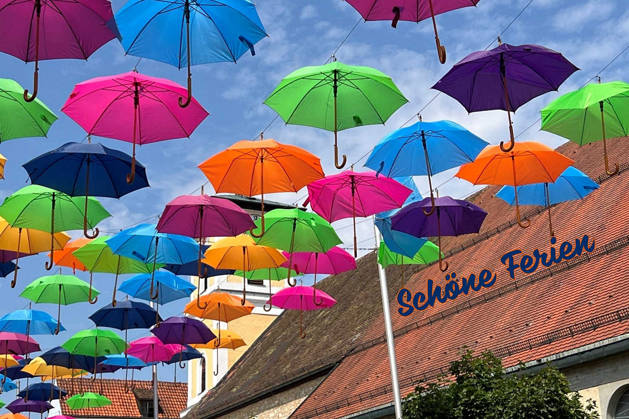 Farbenfrohe Regenschirme über einer Straße und der Schriftzug ‚Schöne Ferien‘ schaffen eine sommerliche Stimmung und wecken V