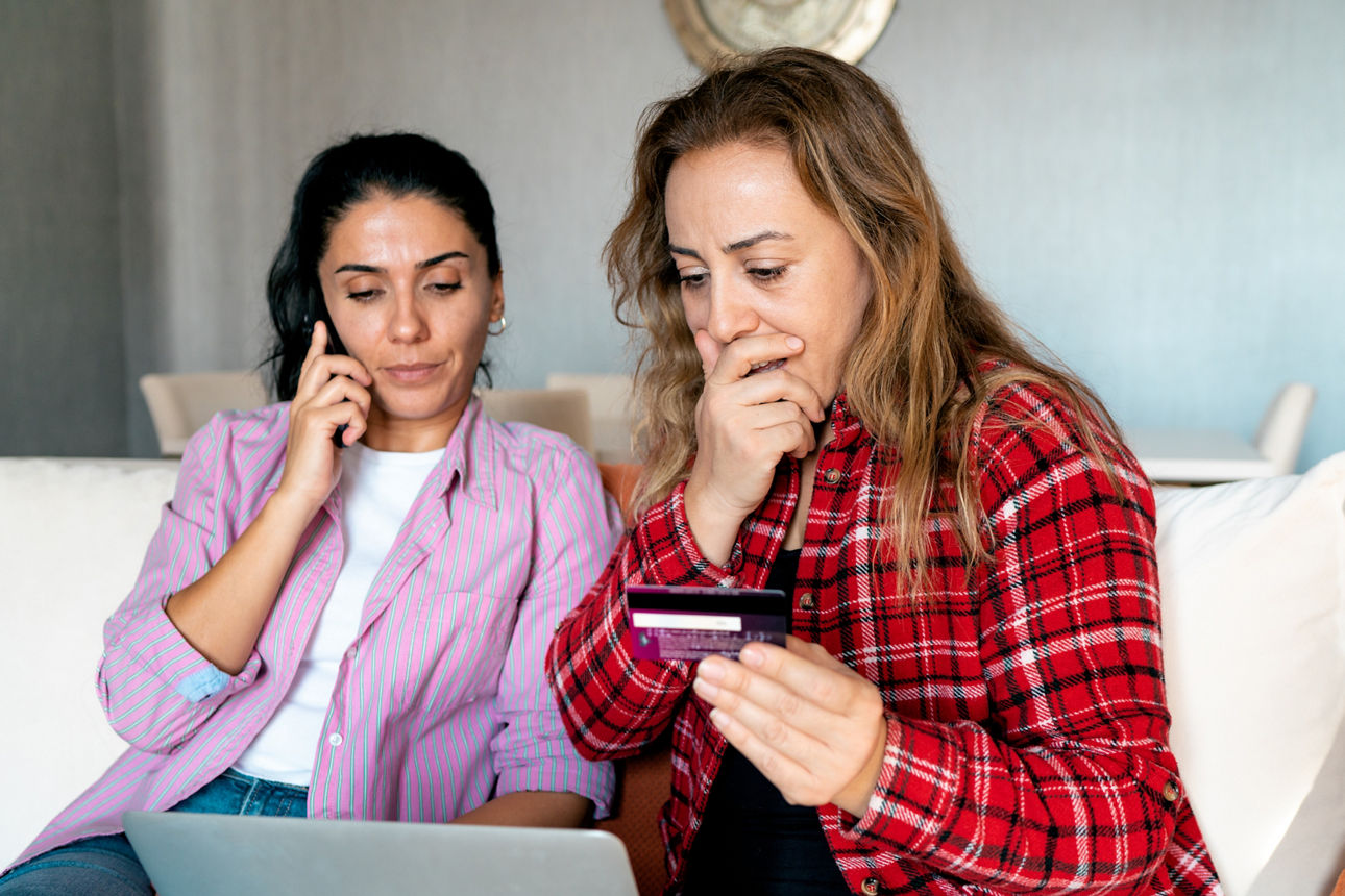 Zwei sitzende, gestresste Frauen halten eine Bankkarte und wirken überrascht.