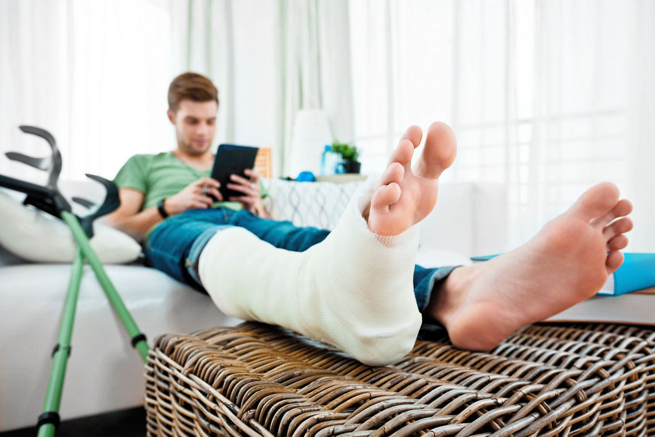 Junger Mann mit gebrochenem Bein sitzt zu Hause auf dem Sofa und liest ein E-Book.