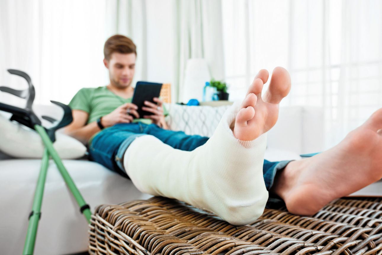 Junger Mann mit gebrochenem Bein sitzt zu Hause auf dem Sofa und liest ein E-Book.