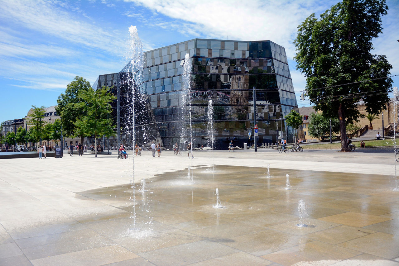 Offener Platz mit Springbrunnen, Bäumen und einem modernen Gebäude mit Spiegelglasfassade.