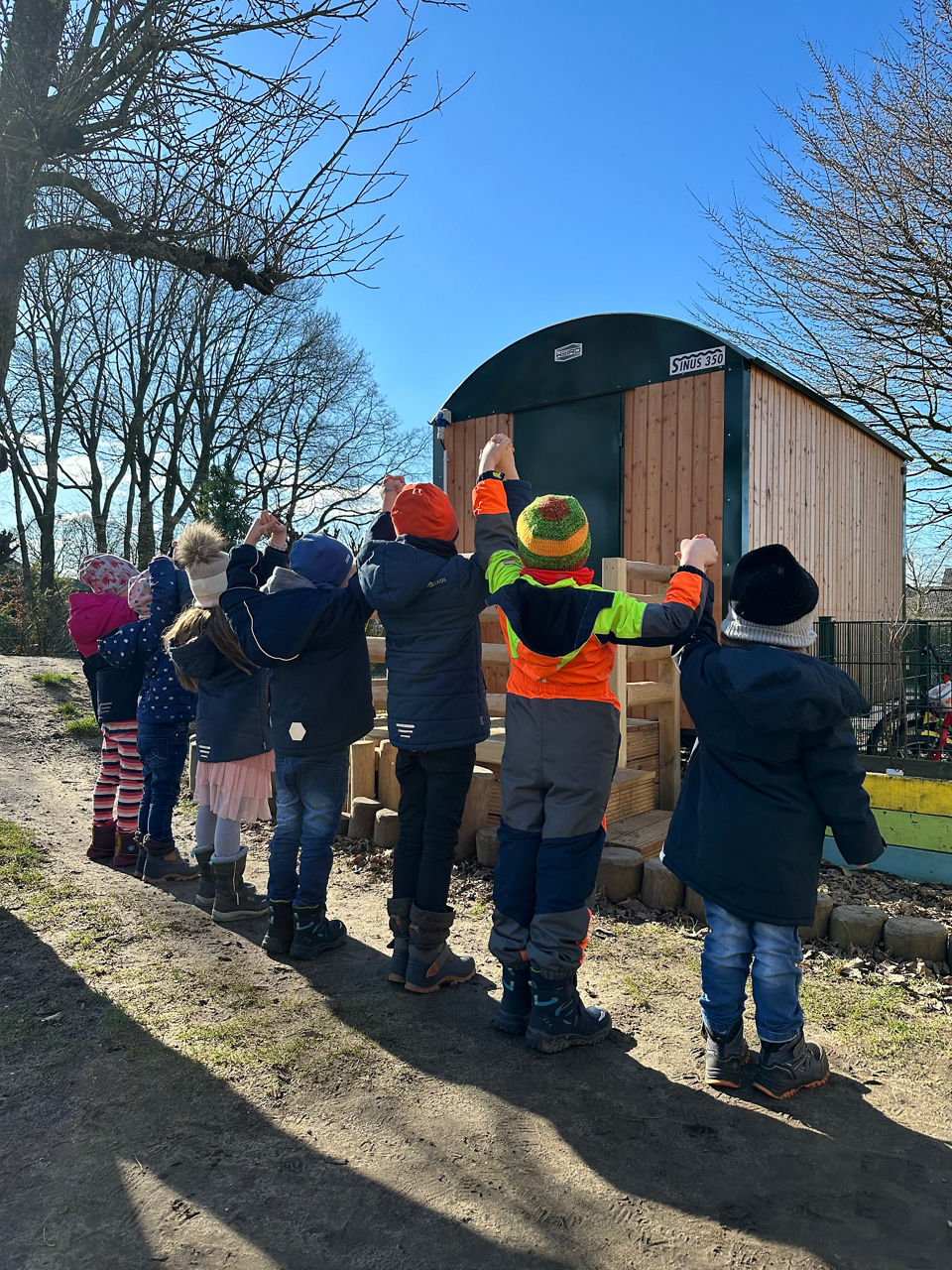 Eine große Gruppe kleiner Kinder auf einer Wiese, neben denen Lehrer und Bankvertreter stehen