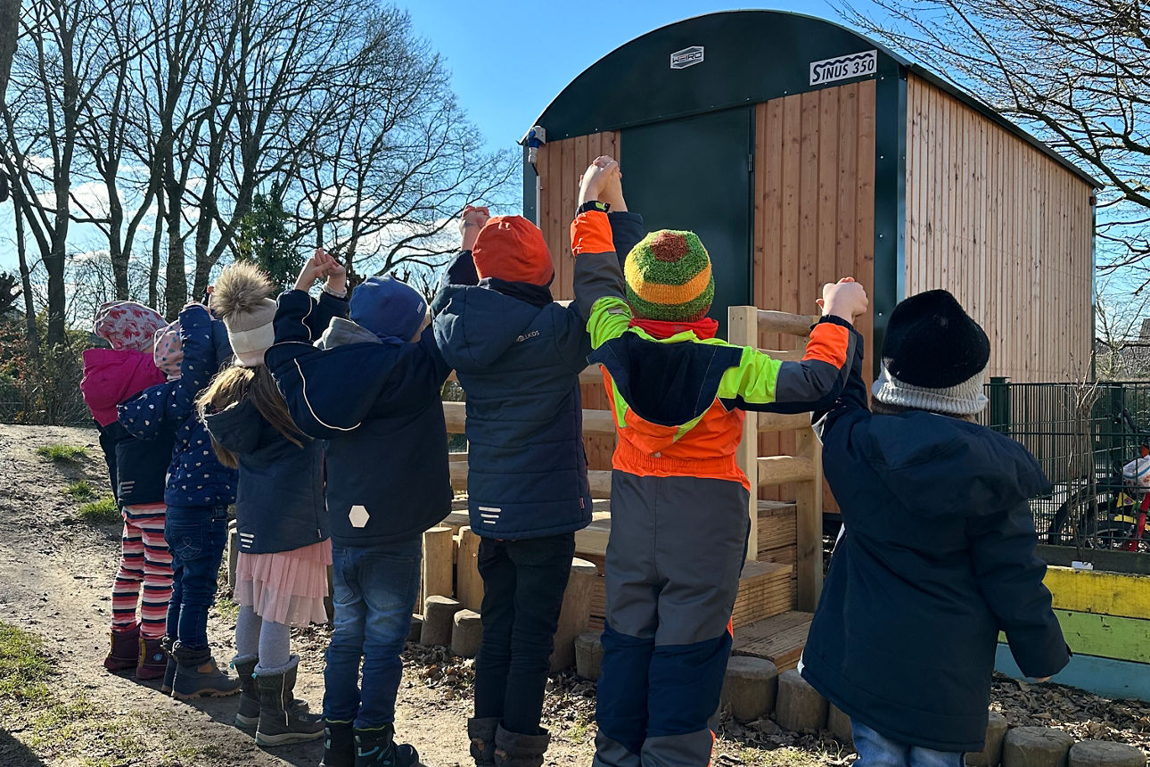 Eine große Gruppe kleiner Kinder auf einer Wiese, neben denen Lehrer und Bankvertreter stehen