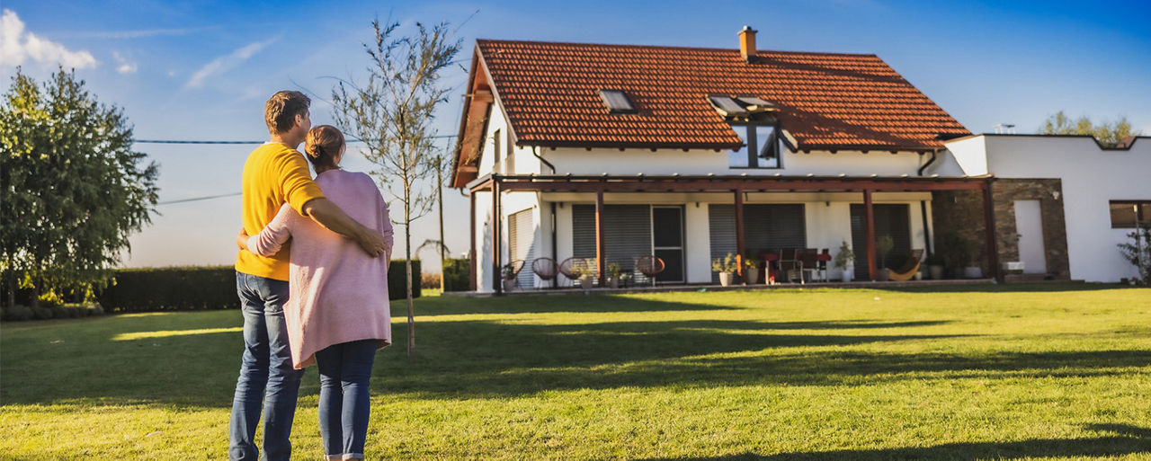 Ein Paar steht vor einem Haus. Sie sind von hinten zu sehen. Sie stehen auf einer großen Wiese vor dem Haus