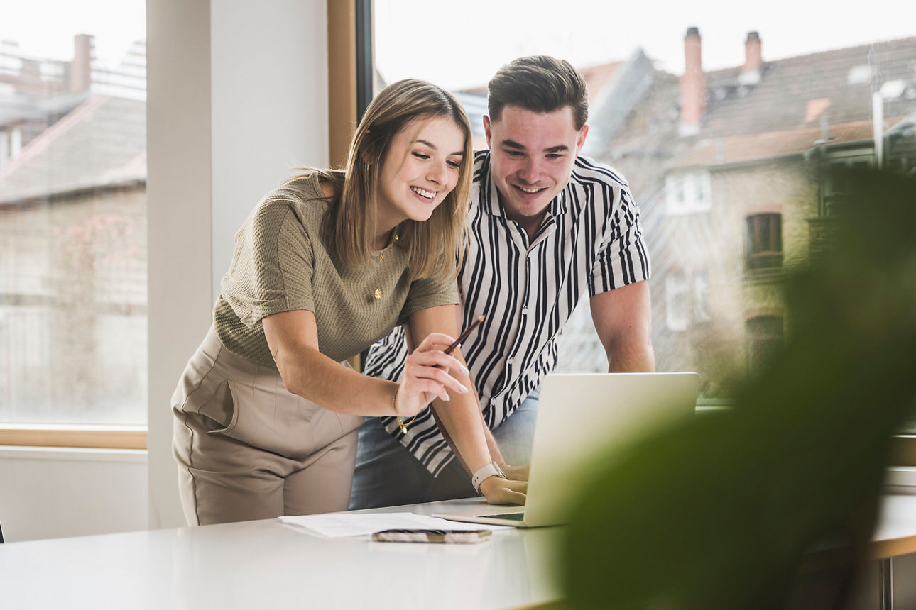 Eine Frau und ein Mann schauen lächelnd auf einen Laptop. Die Frau zeigt em Mann etwas darauf