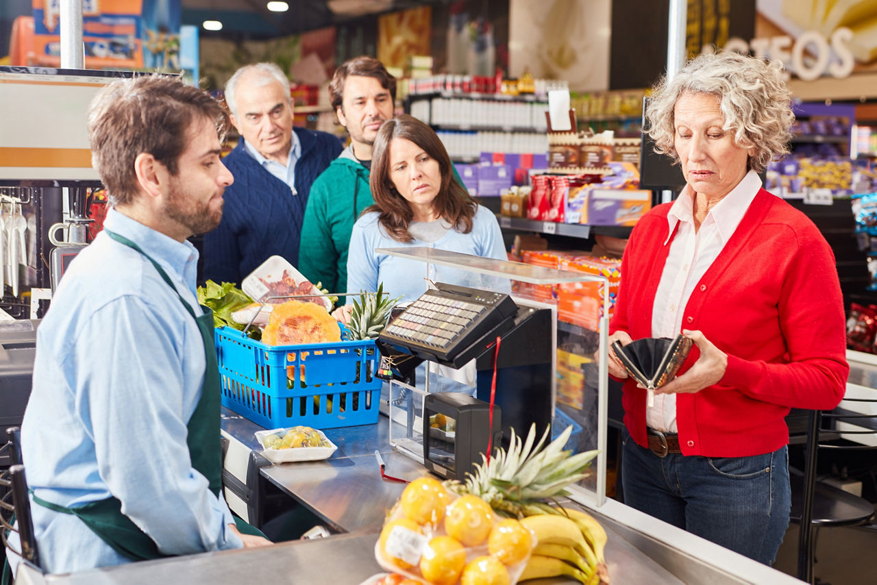 Ältere Frau zahlt mit Bargeld an Supermarktkasse, es bildet sich eine Schlange